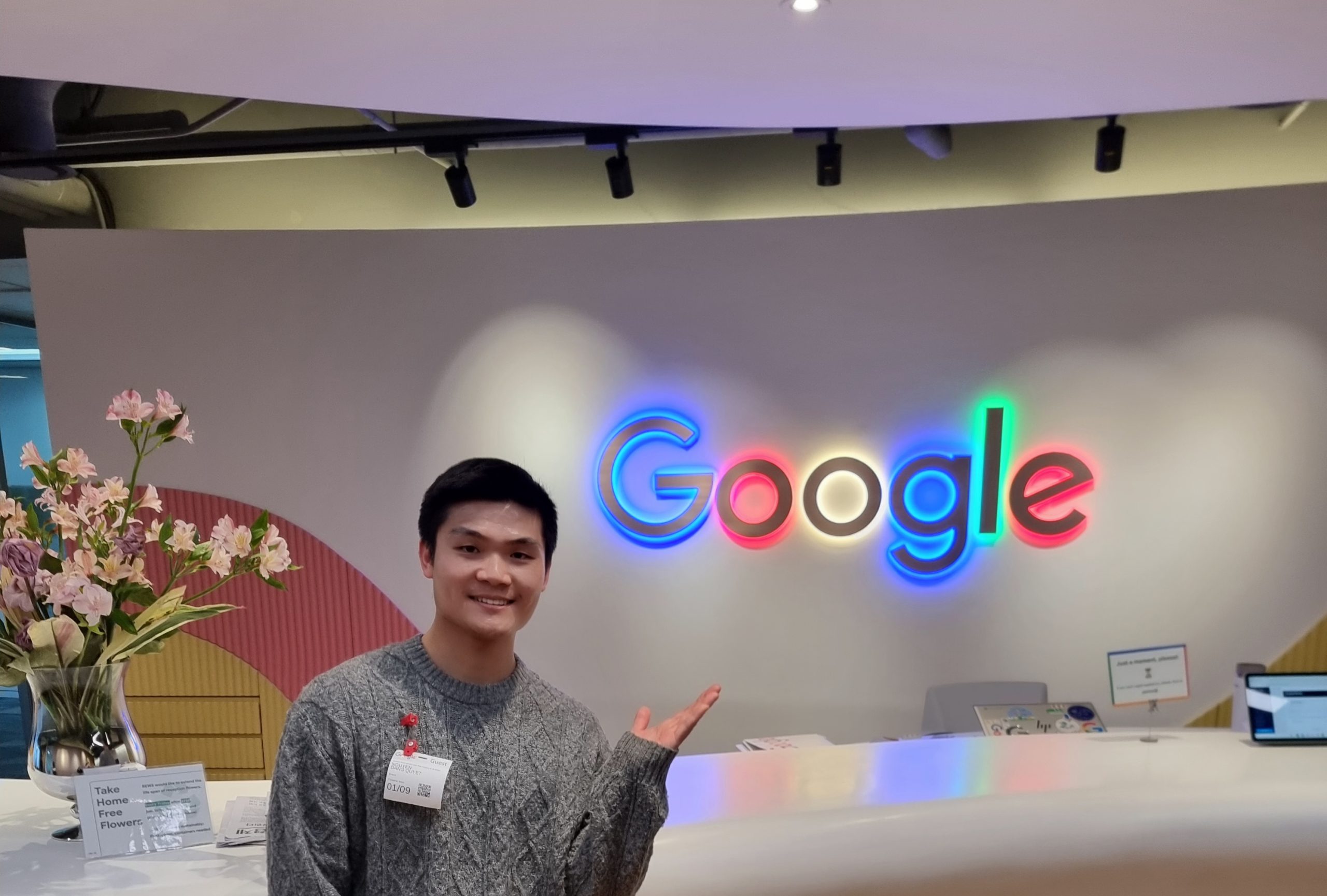 A young Vietnamese international student smiling inside the Google Korea office while pointing toward the illuminated Google logo at the reception area. The modern office interior features colorful branding, soft lighting, and floral decorations, creating a professional and welcoming atmosphere.